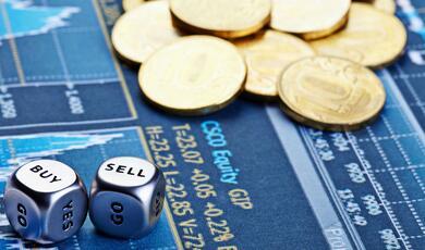 a pile of coins, next to two silvery metal dice with 'buy' and 'sell' on the uppermost faces, on a background of financial data graphs