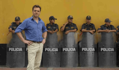 Niall Ferguson standing, hands on hips, in front of a row of police behind riot shields, leaning on a mustard yellow wall.