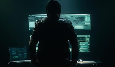 Sillhouette of man in black hoodie in front of multiple glowing computer screens