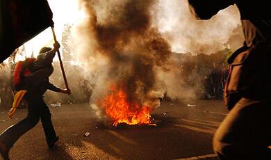 a man waves a flag next to a fire thats burning in the street