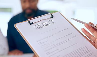 A person holding a "Last Will and Testament" document on a clipboard, with another person out of focus in the background